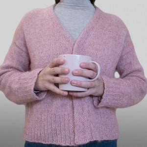 Può includere: Una persona indossa un cardigan di maglia rosa e tiene una tazza di ceramica bianca.