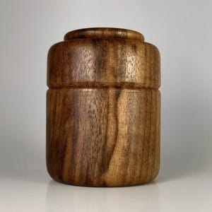 May include: A cylindrical wooden container with a smooth, polished finish and visible dark brown wood grain patterns. The object features a distinct horizontal seam separating the lid from the base, with a rounded top edge and a flat bottom base.