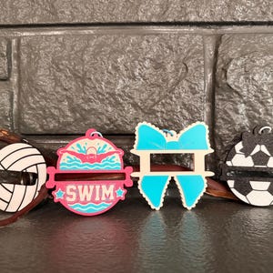 May include: Four decorative acrylic ornaments featuring sports and activity themes. From left to right: a white and black volleyball, a pink and blue circular ornament with a swimmer graphic and the text SWIM, a bright blue bow, and a black and white soccer ball.