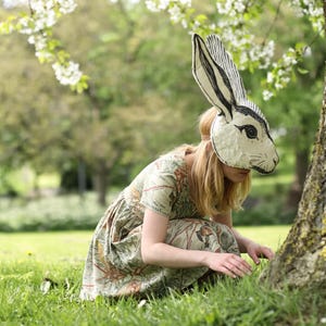 May include: A handmade papier-mâché rabbit mask featuring black ink illustrations of fur and long ears, worn with a short-sleeved dress decorated with a detailed botanical print of green leaves, brown branches, and small orange flowers on a light background.
