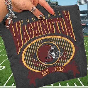 May include: A charcoal gray t-shirt featuring a distressed graphic with the text WASHINGTON FOOTBALL and EST. 1932. The design includes a maroon football helmet inside a yellow oval frame with vertical stripes, rendered in maroon and yellow ink.
