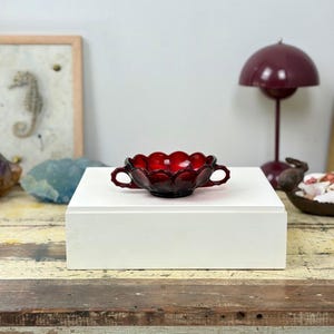 May include: A small, deep red glass bowl featuring a scalloped rim and two side handles. The bowl is crafted from translucent, vibrant red glass with a smooth, reflective surface and a distinct, rounded shape with a wide, open top design.
