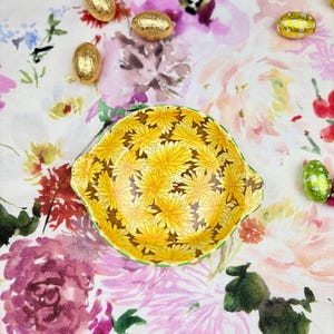 May include: A small, round ceramic bowl featuring a vibrant yellow dandelion pattern on the interior, with a green scalloped rim. The bowl is surrounded by several small, shiny, gold-wrapped chocolate eggs on a floral-patterned fabric surface.