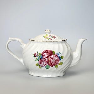 May include: A white ceramic teapot featuring a swirling ribbed texture and a floral pattern of deep pink roses with green leaves and small blue flowers. The teapot includes a matching lid with a small knob and a gold-trimmed rim on the lid.