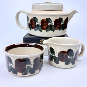 May include: A ceramic tea set featuring a teapot with a lid, a creamer, and a sugar bowl. Each piece is off-white with a repeating pattern of brown circular fruits and dark green leaves encircling the base of the teapot and the sides of the vessels.