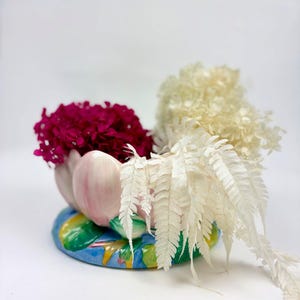 以下が含まれることがあります： 青い土台の上に緑の葉をあしらった、ピンクの花の形をした陶器製のボウル。ボウルの中には、濃いマゼンタ色のアジサイの花の房と、乾燥した白いシダの葉、そしてクリーム色のアジサイの花びらが横にこぼれ落ちるように飾られています。