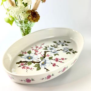 Könnte beinhalten: Eine ovale weiße Keramik-Servierschale mit einer zentralen botanischen Illustration aus hellblauen Blüten mit gelben Mitten und kleinen rosa Blüten an braunen Zweigen. Der äußere Rand ist mit einem wiederkehrenden Muster aus kleinen rosa Blüten verziert.