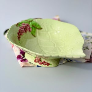May include: A light green ceramic bowl shaped like a leaf with a textured surface and a crackled glaze finish. The bowl features painted pink floral clusters on the rim and base, with green leaves and a raised, pointed edge design.