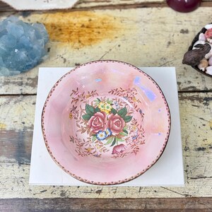 May include: A round, iridescent pink ceramic bowl featuring a central floral motif of pink roses, yellow blossoms, and blue forget-me-nots surrounded by delicate brown foliage. The bowl has a scalloped edge with a dark brown decorative border.