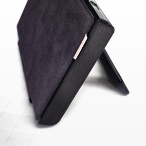 May include: A rectangular electronic device case featuring a dark purple, textured, suede-like front cover and a black, rigid, textured frame. The case is propped open at an angle by an integrated black stand, revealing a thin, light pink edge detail.