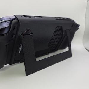 May include: A black handheld gaming console featuring a matte finish, a rear-facing ventilation grille, and a foldable kickstand extended to support the device. The console includes shoulder buttons labeled RT and RB, with a visible serial number sticker.