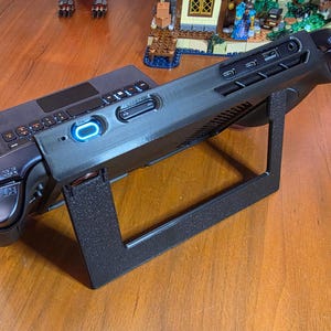 May include: A black handheld gaming console resting on a black textured plastic stand. The device features a glowing blue power button, top-mounted shoulder buttons labeled RT and RB, and various ports including a headphone jack and USB-C input.