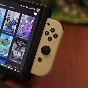 May include: A handheld gaming console featuring a black screen displaying a grid of game titles and a white detachable controller on the right side. The controller includes a black analog stick, a circular home button, and four buttons labeled X, Y, A, and B.
