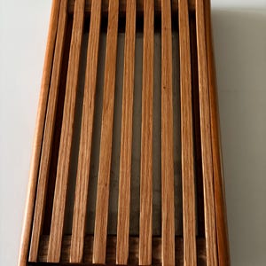 May include: A rectangular wooden bread board featuring a slatted design with evenly spaced vertical slats set within a solid wood frame. The natural light brown wood grain is visible throughout the structure, which has a flat, open-top surface.