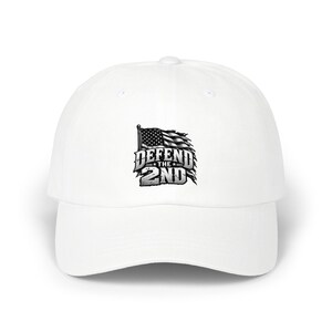 May include: A white baseball cap featuring a graphic on the front panel. The graphic displays a distressed, black and white American flag waving above the bold, metallic-style text that reads DEFEND THE 2ND in a stylized, blocky font.