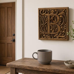 May include: A square wooden wall plaque featuring a carved letter B surrounded by intricate scrollwork patterns. The plaque is mounted on a plain wall above a wooden table that holds a grey ribbed mug, a small bowl with keys, a vase, and a candle.