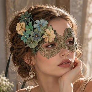 Peut inclure: Un masque de carnaval en dentelle vert olive orné d'un motif en filigrane complexe. Le masque est décoré d'un groupe de fleurs en tissu dans des tons bleu doux, jaune pâle et vert sauge, chacune dotée d'une petite perle centrale.