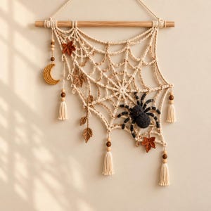 Puede incluir: Un tapiz de macramé color crema con forma de telaraña, colgado de un listón de madera. Incluye una araña tejida en gris oscuro, una luna creciente tejida en amarillo dorado y hojas marrones, decorado con cuentas de madera y borlas colgantes.