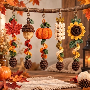 Può includere: Cinque ornamenti pendenti all'uncinetto appesi a un ramo, raffiguranti una foglia d'acero rossa, una ghianda marrone, una zucca arancione, un grappolo floreale bianco e un girasole giallo, ciascuno con un viticcio a spirale e una pigna.
