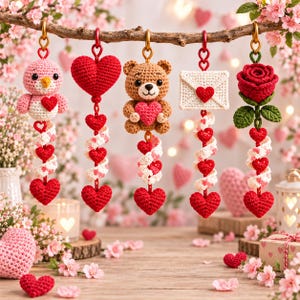 May include: Five crocheted hanging ornaments suspended from a branch, featuring a pink bird, a red heart, a brown teddy bear, a white envelope, and a red rose. Each ornament includes a spiral of small red and white hearts dangling below.
