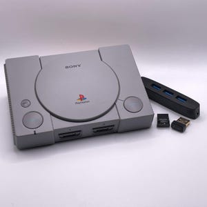 May include: A grey Sony PlayStation games console featuring a circular disc cover with the PlayStation logo, two controller ports, and power and reset buttons. Included are a black USB hub with three ports and two small black USB flash drives.