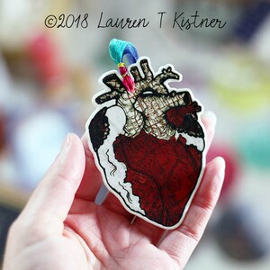 May include: A red and white anatomical heart ornament with a plaid pattern. The ornament is hanging from a blue and green ribbon.