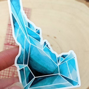 Puede incluir: Una pegatina de vinilo brillante en forma de cristal azul y turquesa del estado de Idaho. La pegatina tiene un borde blanco.