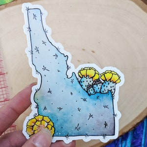 Op de afbeelding: Een blauwe en grijze waterverf sticker van de staat Idaho met een cactusontwerp en gele bloemen. De sticker is zwart omlijnd en heeft een witte achtergrond.