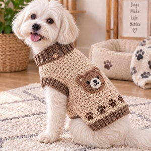 May include: A beige crocheted dog sweater featuring a dark brown ribbed collar and hem. The sweater is decorated with a brown bear face appliqué on the side and a row of dark brown paw print motifs along the bottom edge. A small wooden button is visible.