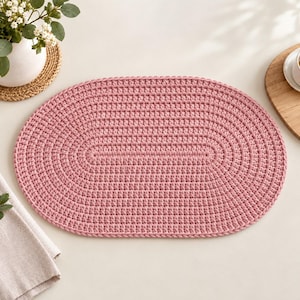 May include: An oval-shaped, dusty pink crocheted placemat featuring a consistent, textured stitch pattern throughout. The placemat is constructed with a dense, uniform weave and finished with a clean, braided edge along the entire perimeter.
