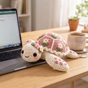 Puede incluir: Un peluche de tortuga tejido a ganchillo con cabeza y extremidades de color crema con detalles en rosa. El caparazón presenta un patrón de parches hexagonales rosas, cada uno con una margarita blanca con hojas verdes y un borde festoneado.