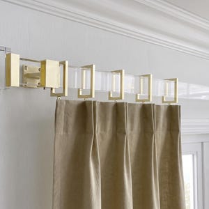 May include: A clear acrylic curtain rod featuring brushed gold metal brackets and square-shaped curtain rings. The rod is mounted to the wall and holds pleated beige fabric curtains, showcasing a clean, structural design with metallic accents.
