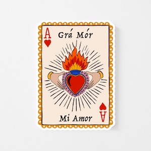 May include: A sticker featuring a playing card design with a heart-shaped emblem. The card is beige with a gold border and the text "Grá Mór" and "Mi Amor". The central image is a heart with flames, held by two hands.