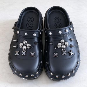May include: A pair of black slip-on clogs featuring silver-toned metal embellishments. The shoes are decorated with circular studs along the perimeter, sharp spikes, clusters of small skull charms, and metallic X-shaped accents on the top surface.