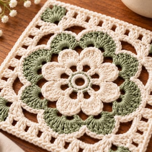 May include: A square crochet coaster featuring a central cream-colored flower motif surrounded by sage green fan-shaped stitches. The intricate needlework creates a geometric pattern with open spaces, finished with a cream-colored border.