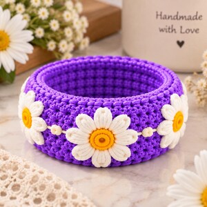 May include: A circular purple crochet bangle bracelet featuring three-dimensional white daisy flowers with bright yellow centers attached around the exterior. The bracelet has a thick, textured, woven yarn construction with a consistent pattern.