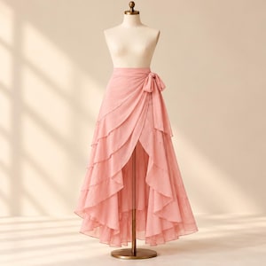 May include: A dusty pink wrap skirt displayed on a mannequin. The skirt features multiple cascading ruffled layers that create a high-low hemline, with a fabric tie closure at the waist. The material appears lightweight and semi-sheer.