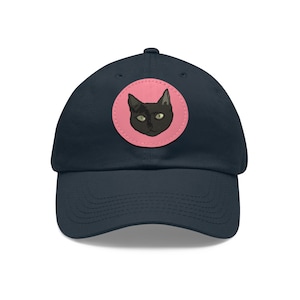 May include: A navy blue baseball cap with a pink circle patch featuring a black cat with green eyes.