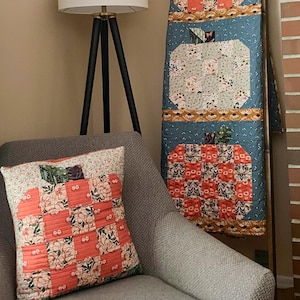 Puede incluir: Una colcha rectangular y un cojín cuadrado a juego con un diseño de calabaza de patchwork. La colcha y el cojín están confeccionados con telas en tonos coral, azul y crema, con estampados florales y puntadas de acolchado horizontales visibles.