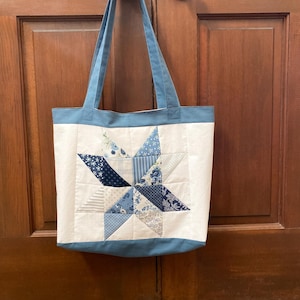 May include: A white tote bag featuring a blue star quilt design. The star is constructed from various blue patterned fabrics. The bag has blue handles and a blue border. The bag is suspended on a wooden door.