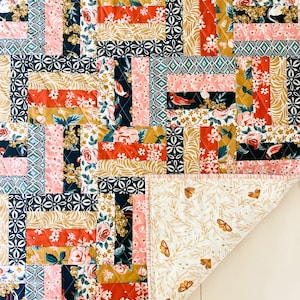 May include: A patchwork quilt with a mix of floral and geometric patterns in pink, coral, navy, gold, and white. The quilt's design incorporates various fabrics and textures. The backing is white, decorated with gold butterflies and leaves.