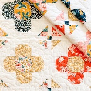 Puede incluir: Un edredón hecho a mano con un diseño de patchwork con patrones florales y geométricos. El edredón muestra una variedad de colores, incluyendo blanco, rosa, dorado, rojo y azul marino. El diseño incluye bloques en forma de cruz y acentos triangulares, con una textura acolchada.