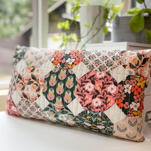 May include: Rectangular decorative pillow with a patchwork design. The pillow features a variety of floral patterns in shades of pink, orange, green, and cream. The pillow is placed on a white surface near a window with plants.
