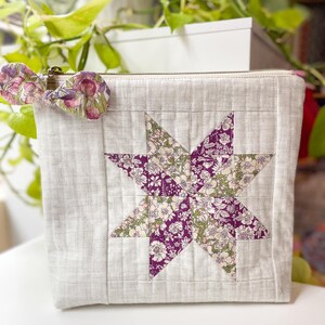 May include: A light grey quilted zip-up pouch featuring a patchwork star design. The star is made from floral fabric in purple, green, and white. A matching floral bow is attached to the zip. The pouch is on a white surface.
