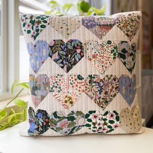 Puede incluir: Un cojín decorativo cuadrado con un patrón de corazones acolchados. Los corazones están hechos de varias telas florales en tonos de rosa, azul, verde y crema. El fondo es de color crema sólido, y el cojín está sobre una superficie blanca.