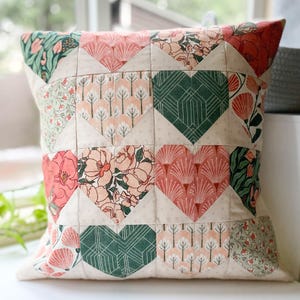 Puede incluir: Cojín decorativo cuadrado con diseño de patchwork. El cojín presenta patrones en forma de corazón en varias estampados florales y geométricos, incluyendo colores rosa, verde y crema. El cojín es probablemente para la decoración del hogar.