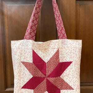 May include: A quilted tote bag with a large, eight-pointed star design in shades of red. The star is centered on a cream-colored background with a delicate floral pattern. The handles are a coordinating red floral print.