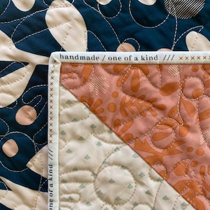 May include: Close-up of a handmade quilt with a navy blue and cream floral pattern. The quilt features a diagonal section with a peach and cream design. The edge of the quilt has the text "handmade / one of a kind".