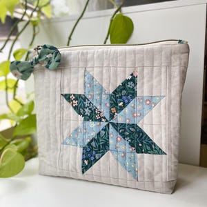 May include: A beige quilted pouch featuring a starburst design in blue and green floral patterns. The pouch has a zip closure and a small fabric bow attached. The bag is made of quilted fabric.