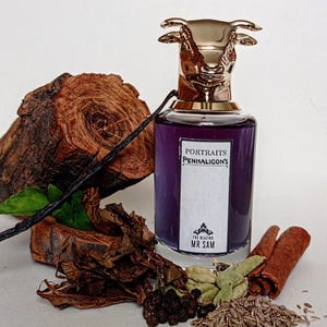 May include: A glass perfume bottle filled with purple liquid, topped with a gold-colored bull head cap. The label reads Portraits Penhaligon's The Blazing Mr Sam. It is surrounded by a wood log, a vanilla bean, tobacco leaves, cardamom, peppercorns, and cinnamon.